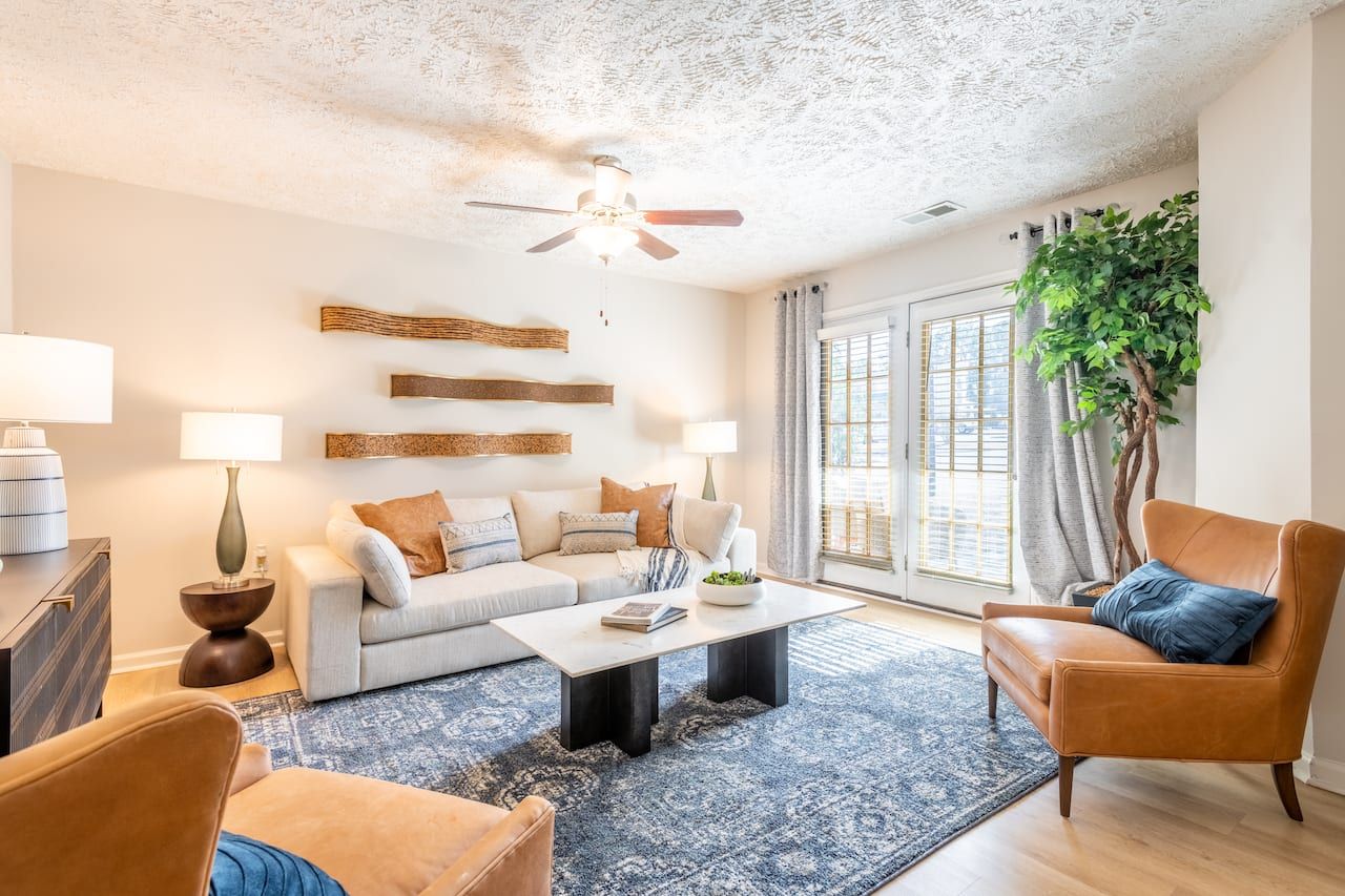 Bright living room with a white sofa, brown chairs, blue rug, wall art, and large windows with curtains.
