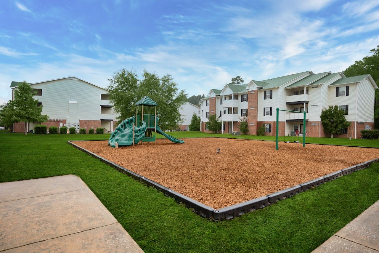 Playground with slides and swings surrounded by grass, near multi-story apartment buildings on a clear day.
