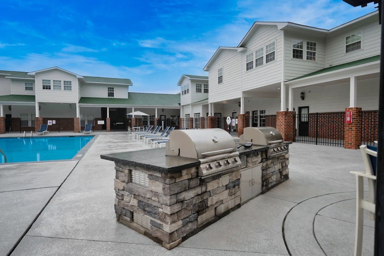 Outdoor pool area with lounge chairs and dual grills, surrounded by apartment buildings under a blue sky.